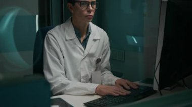 Vitrinin arkasından, MRI makinesinin yansımasıyla, bilgisayarla yazı yazan ve zor durumdaki hasta hakkında bilgi toplayan olgun beyaz kadın radyografın orta şiddette görüntüsü.