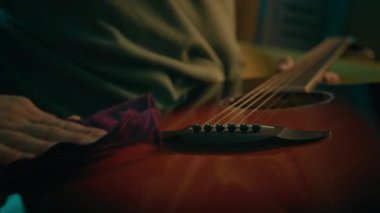 Akustik gitarla ilgilenen, neon ışıklı odada yumuşak havluyla enstrümanın yüzeyini temizleyen isimsiz müzisyenlerin yakın plan çekimleri.