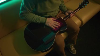 Neon ışıklı odada leaher koltukta otururken akustik gitar gövdesini yumuşak bir bezle temizleyen bir adamın yukarıdan aşağı görüntüsü.