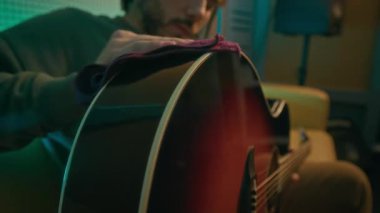 Akustik gitarla ilgilenen, neon ışıklı odada yumuşak bir bezle yüzeyini temizleyen bir adamın yakın plan çekimleri.