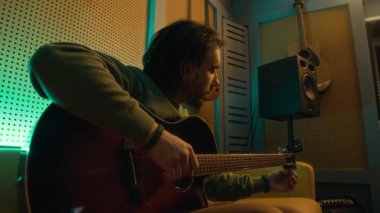 Erkek müzisyenin kanepeye oturup akustik gitar akort ederken loş neon ışıklı stüdyoda elektronik akort cihazı kullanırken ki yan görüntüsü.