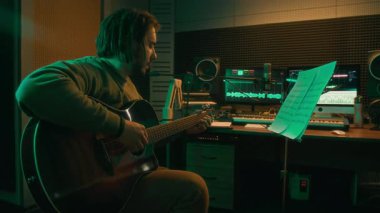 Beyaz erkek müzisyenin akustik gitar tıngırdattığı, arka planda bilgisayar monitörleri ve ekipmanlarıyla neon ışıklı kayıt stüdyosundaki notalardan yeni şarkı ya da melodi öğrendiği yan çekim.