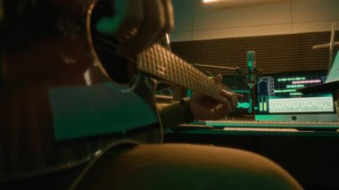 Akustik gitar tıngırdatan, arka planda bilgisayar monitörleriyle neon ışıklı kayıt stüdyosunda şarkı çalan isimsiz gitaristin yakın plan çekimleri.