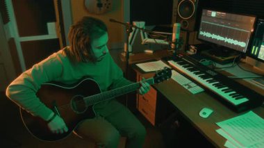 Gitar çalan adamın yüksek açılı görüntüsü veya yeşil neon ışıklı stüdyodaki bilgisayara gitar sesi kaydetmesi, arka plandaki bilgisayar monitöründeki ses dizisi