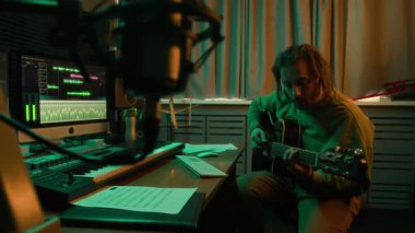 Sakallı, uzun saçlı bir adamın gitar çalıp, kaydedilmiş ses sekanslarıyla bilgisayar programına ses kaydederken, profesyonel ekipmanlarla modern yeşil ışıklı stüdyoda yeni bir müzik yaratırken görüntüsü.