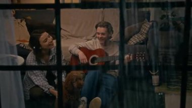 Tam kare, pencereden bakıldığında, evcil köpekli genç beyaz bir çift yağmurlu bir gecede evde, salonda oturmuş gitar çalan bir adam ve şarkı söyleyen bir kadın.
