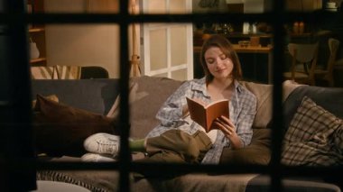 Tam kare, sokaktan pencereye bakarken, rahat bir dairede akşamları evde yalnız kalan, kanepede heyecanlı kitaplar okuyan, gülümseyen, sayfaları karıştıran beyaz bir kadın.