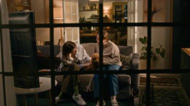 Neşeli genç beyaz çiftin, erkek arkadaşın ve kız arkadaşının pencerelerinden içeri girip kanepede oturup, evcil maltipoo köpeğiyle oynayarak, tüylü köknarını okşayarak, kahkahalarla...