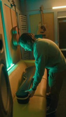 Kafkasyalı erkek müzisyenin, gitarı neon ışıklı kayıt stüdyosuna getirirken tam dikey görüntüsü.
