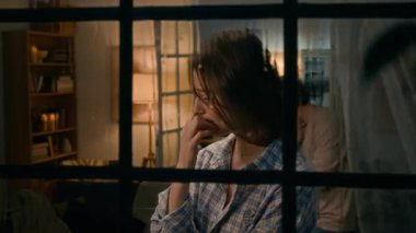 Akşamki fırtına ve yağmur sırasında pencerenin kenarında duran ağlayan ve hıçkıra hıçkıra ağlayan ağlayan ağlayan, arka planda volta atan ağlayan genç beyaz kadının orta boy yakın çekimi.