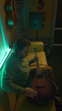 Kafkasyalı erkek gitaristin kanepede oturup, neon ışıklı kayıt stüdyosundaki yumuşak havluyla gitar silerken, enstrümanla ilgilenirken dikey yan görüntüsü.