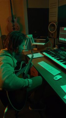 Gitar çalan erkek sanatçının dikey yan görüntüsü modern yeşil ışıklı stüdyoda daha fazla düzenleme yapmak için sesini bilgisayara kaydediyor.