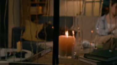 Yağmurlu bir gecede yanan mum dolu bir tablo ve battaniyeyle kanepede oturan genç bir kadın, yabancı ya da sapık tarafından pencereden görülen bulanık arka planda kitap okuyor.