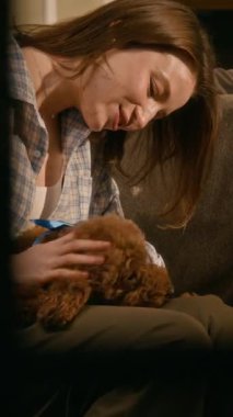 Pencerede görülen orta boy dikey fotoğraf, keyifli beyaz bir kızın koltukta oturup, mavi kurdeleli evcil bir maltipoo köpeğiyle oynaşması.