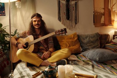 Mumlarla, ev bitkileriyle ve sanatsal dekorlarla çevrili sıcacık bir bohem tarzı odada renkli yastıklarla rahat bir yatakta otururken müziğin tadını çıkarmak.
