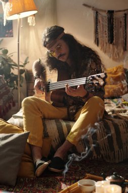 Uzun saçlı ve güneş gözlüklü bir adam rahat, bohem esinlenilmiş bir oturma odasında gitar çalıyor. Bitkiler ve hafif ışıklandırmayla dolu. Rahat bir atmosfer yaratıyor.