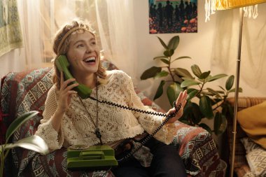 Kafkasyalı bir kadın yeşil bir telefonu tutarken gülüyordu rahat, bitkilerle dolu bir oturma odasında oturuyordu. Gün ışığı hafifçe uzayı aydınlatıyordu.