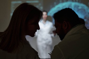 Tıbbi konferans sırasında seyircilere fısıldayan iki kişinin silueti, boşluğu kopyala