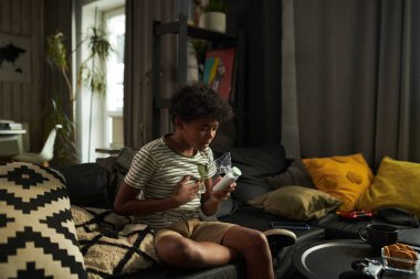 Göğsünde yastıkla kanepede oturan ve oksijen maskesiyle astım spreyine bakan genç Afrikalı Amerikalı çocuk.