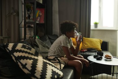 Astımlı genç Afro-Amerikalı çocuk televizyon izlerken kanepede oturup sprey maskesinden oksijen soluyor.