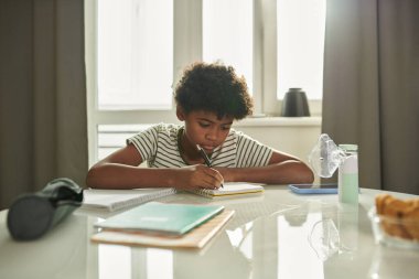 Okula gitmeden önce masayı eğip ödevlerini yaparken not alan tatlı öğrenci.