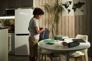 Astımlı bir çocuğun kahvaltıdan sonra mutfak masasının yanında okul eşyalarını sırt çantasına koyuşunun yan görüntüsü.