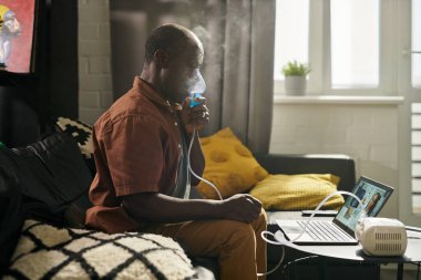 Laptopun önünde oturan ve sanal sağlık asistanına danışan olgun bir adamın nebulizatörden buhar soluyuşunun yan görüntüsü