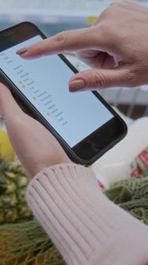 Alışveriş listesiyle akıllı telefonu tutan ve markette ekranı dinleyen tanınmayan bir kadının dikey görüntüsü.