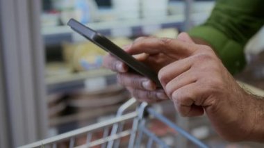 Süpermarkette akıllı telefon kullanan ve ekran kontrol listesini araklayan tanınmayan bir adamın yakın plan görüntüsü.