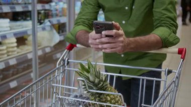 Süpermarkette akıllı telefon kullanan, alışveriş arabasına yaslanmış, alışveriş listesini kontrol eden, ekranı kopyalayan, uzayı kopyalayan, tanınamayan bir adamı yakından çek.