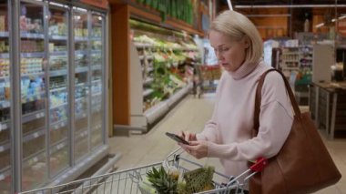 Markette akıllı telefon kullanan sarışın bir kadının alışveriş arabasına yaslanıp alışveriş listesine bakarken çekilmiş yan görüntüsü.