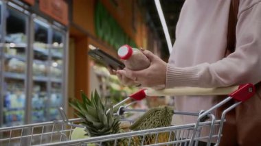 Süpermarkette akıllı telefon kullanan, süt ürünlerindeki qr kodlarını tarayan ya da malzeme listesini kontrol eden tanınmayan yetişkin bir kadının yan görüntüsü.