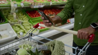 Süpermarkette alışveriş arabasını iterken akıllı telefon kullanan tanınmayan bir yetişkinin yakın plan fotoğrafı, boşluğu kopyala