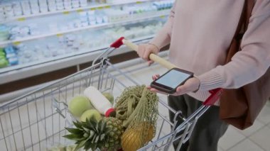 Yüksek açılı, tanınmayan bir kadının alışveriş listesi ile akıllı telefonu tutarken, süpermarkette el arabası iterken, mekanı kopyaladığı yakın çekim.