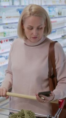 Süpermarkette alışveriş yaparken gülümseyen ve elinde telefon tutan sarışın kadının dikey görüntüsü.