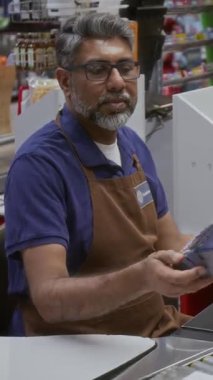 Dükkanda çalışan yetişkin bir adamın dikey çekimi ve bakkaliye alışverişi yapan müşterilere banka terminali vermek.