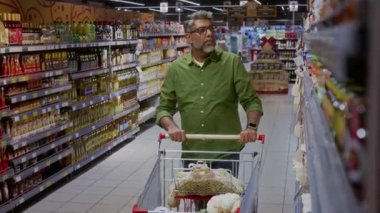Olgun Hintli adam süpermarkette alışveriş arabasını kameraya doğru itiyor ve yiyecekleri seçerken üst raftan bir şeyler alıyor.