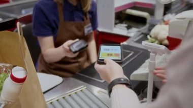 Yüksek açılı yakın çekim, tanınmayan kadın müşterinin süpermarketteki kasada ödeme yaptığı ve ekranda indirim kodu olan akıllı telefonu kasiyere verdiği.