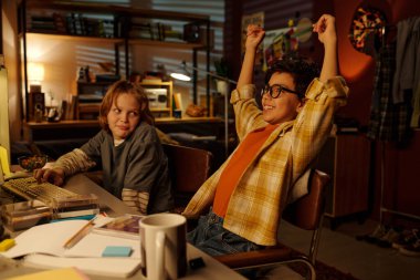 Yatak odasında oynayan iki çocuk, oyun zaferini kutlarken kollar havada ve neşeli ifadelerle, kitaplar, defterler ve bilgisayar ekipmanlarıyla çevrili.