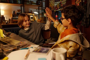 İki çocuk çalışma odasında oturmuş çeşitli kitaplar ve çalışma materyalleriyle çevrili ortak bir başarı için beşlik çakıyor ve verimli ve coşkulu bir atmosfer yaratıyorlar.