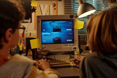 İki kişi karmaşık bir bilgisayar masasında, ekranın etrafında çeşitli notlar ve öğelerle retro oyun oturumuna katılıyor.