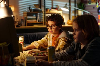 İki çocuk, biri gözlüklü, loş ışıklı odada bilgisayar oyunu oynuyorlar. İçi çeşitli nesneler ve masa lambasından gelen yumuşak bir ışık var.