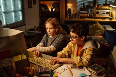 Samimi bir odada raflar ve kitaplarla bilgisayar projesinde çalışan iki genç arkadaş. Akşam ışığı çalışma alanını aydınlatıyor ve sıcak bir atmosfer yaratıyor.