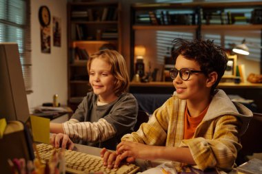 Masanın etrafında kitap ve kişisel eşyalarla dolu sıcak bir odada bilgisayarla gülümseyen iki çocuk rahat bir ortam yaratıyor.