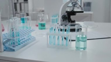 Laboratuvar masasında açık mavi çözeltisi, arka planda mikroskop olan raflarda cam deney tüpü ve birden fazla test tüpü olan insanlar yok.