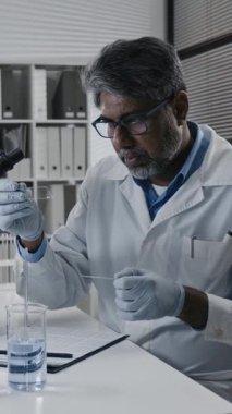 Üst düzey Hintli bilim adamının modern ilaç geliştirme laboratuvarında çalışırken cam numunelerini analiz ederken ve dokümanlara not alırken dikey yan görüntüsü.