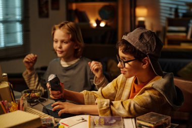 İki çocuk bir masada oturmuş, sıcak aydınlatılmış bir odada video oyunları oynuyorlar, birlikte vakit geçiriyorlar. Kitaplarla dolu raflar ve arka planda bir küre var.