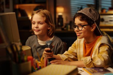 İki küçük çocuk, sıcak ışıklarla dolu çalışma odasındaki renkli kırtasiye malzemelerinin yer aldığı akşam aktivitelerine dalmış bir masada oturuyorlar.