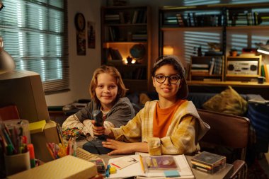 Kitap, dekorasyon ve çalışma malzemeleriyle dolu rahat bir odada video oyunundan hoşlanan iki çocuk, paylaşılan aktivitelere odaklanmış sıcak ve davetkar bir ortam yaratıyor.