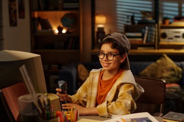 Gülümseyen, gözlüklü ve şapkalı bir çocuk, elinde telsizle rahat, aydınlık bir odada otururken arka planda kitap rafları ve diğer objelerle